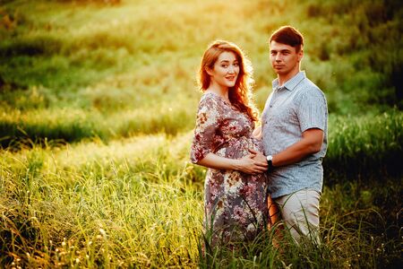 Young happy beautiful couple in love walking together on grass and trees park landscape on sunset in summer with man touching pregnant belly of his woman. Family conceptの写真素材