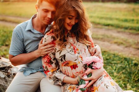 Stylish pregnant couple holding hands on belly in sunny light in summer park. Happy young parents, mom and dad, hugging baby bump and smiling, enjoying beautiful moment at sunset among grassの写真素材