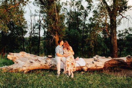 Stylish pregnant couple holding hands on belly in sunny light in summer park. Happy young parents, mom and dad, hugging baby bump and smiling, enjoying beautiful moment at sunset among grassの写真素材