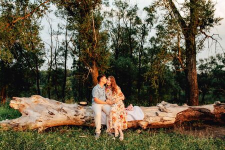 Stylish pregnant couple holding hands on belly in sunny light in summer park. Happy young parents, mom and dad, hugging baby bump and smiling, enjoying beautiful moment at sunset among grassの写真素材