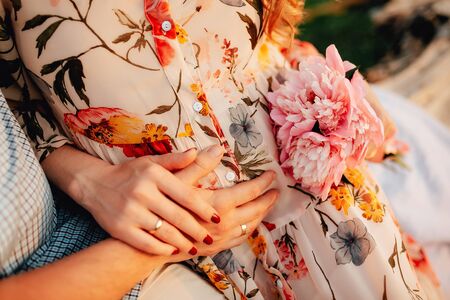 Stylish pregnant couple holding hands on belly in sunny light in summer park. Happy young parents, mom and dad, hugging baby bump and smiling, enjoying beautiful moment at sunset among grassの写真素材