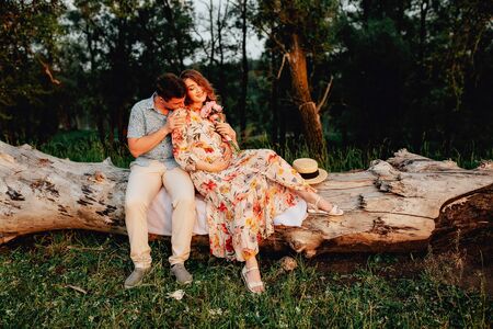 Stylish pregnant couple holding hands on belly in sunny light in summer park. Happy young parents, mom and dad, hugging baby bump and smiling, enjoying beautiful moment at sunset among grassの写真素材