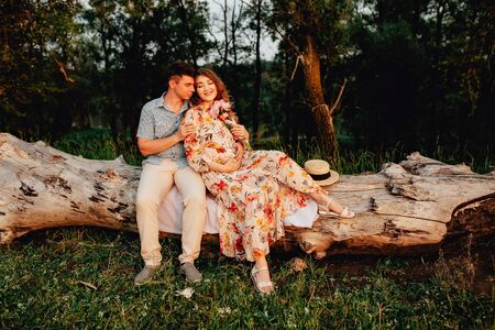 Stylish pregnant couple holding hands on belly in sunny light in summer park. Happy young parents, mom and dad, hugging baby bump and smiling, enjoying beautiful moment at sunset among grassの写真素材