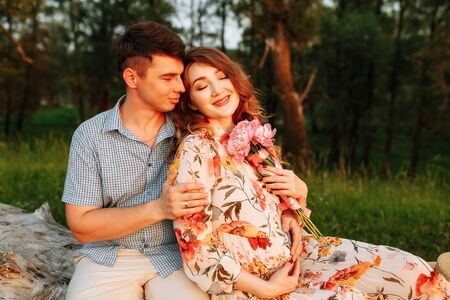 Stylish pregnant couple holding hands on belly in sunny light in summer park. Happy young parents,...