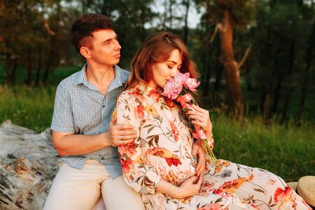 Stylish pregnant couple holding hands on belly in sunny light in summer park. Happy young parents, mom and dad, hugging baby bump and smiling, enjoying beautiful moment at sunset among grassの写真素材