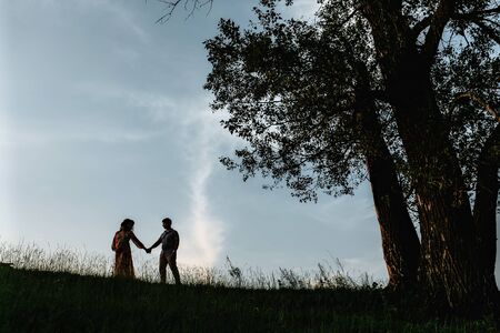 Stylish pregnant couple silhouettes in sunny light in park among grass. Happy young parents, mom and dad, enjoying beautiful moment at sunset. Parenthood conceptの写真素材