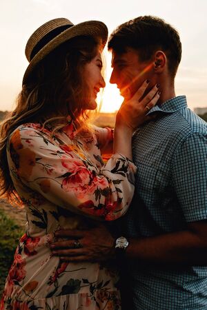 Stylish pregnant couple holding hands on belly in sunny light in summer park. Happy young parents, mom and dad, hugging baby bump and smiling, enjoying beautiful moment at sunset among grassの写真素材
