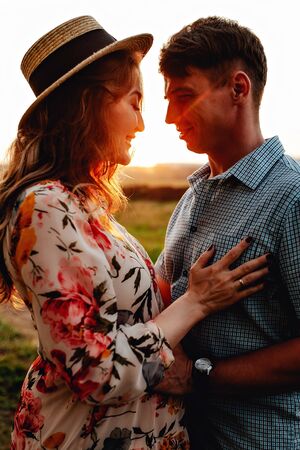 Stylish pregnant couple holding hands on belly in sunny light in summer park. Happy young parents, mom and dad, hugging baby bump and smiling, enjoying beautiful moment at sunset among grassの写真素材