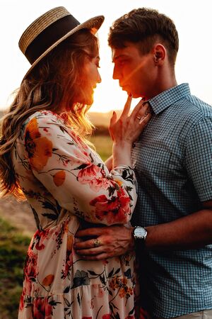Stylish pregnant couple holding hands on belly in sunny light in summer park. Happy young parents, mom and dad, hugging baby bump and smiling, enjoying beautiful moment at sunset among grassの写真素材