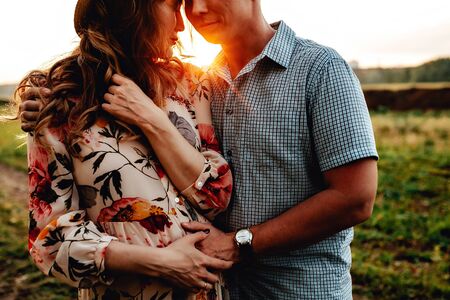 Happy young parents, mom and dad, hugging baby bump and smiling, enjoying beautiful moment at sunset among grass. Stylish pregnant couple holding hands on belly in sunny light in summer park.の写真素材