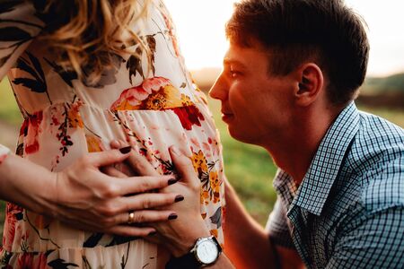 Stylish pregnant couple holding hands on belly in sunny light in summer park. Happy young parents, mom and dad, hugging baby bump and smiling, enjoying beautiful moment at sunset among grassの写真素材