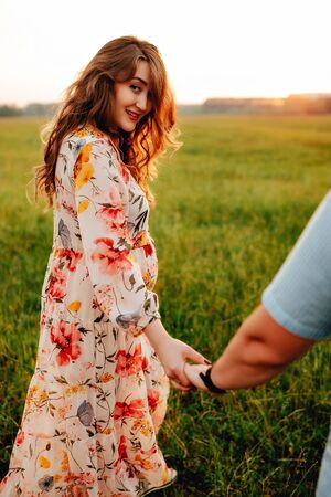 Young happy beautiful couple in love walking together on grass and trees park landscape on sunset in summer with man touching pregnant belly of his woman. Family conceptの写真素材