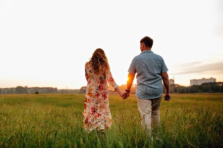 Stylish pregnant couple holding hands on belly in sunny light in summer park. Happy young parents, mom and dad, hugging baby bump and smiling, enjoying beautiful moment at sunset among grassの写真素材
