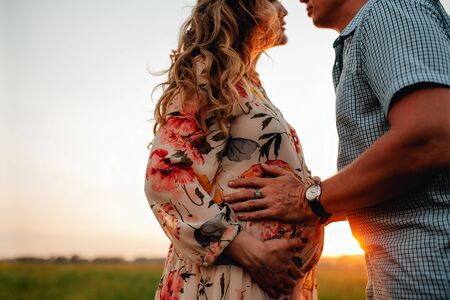 Stylish pregnant couple holding hands on belly in sunny light in summer park. Happy young parents, mom and dad, hugging baby bump and smiling, enjoying beautiful moment at sunset among grassの写真素材