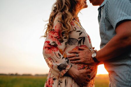 Young happy beautiful couple in love walking together on grass and trees park landscape on sunset in summer with man touching pregnant belly of his woman. Family conceptの写真素材