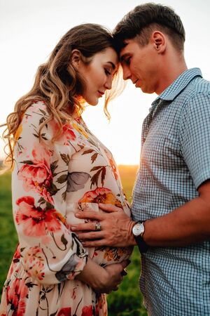 Stylish pregnant couple holding hands on belly in sunny light in summer park. Happy young parents, mom and dad, hugging baby bump and smiling, enjoying beautiful moment at sunset among grassの写真素材