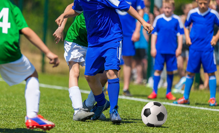 Competition between two youth soccer teams. Boys in blue and green sports uniforms running and kicking a soccer ball on the pitch.の写真素材