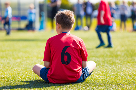 Child dressed in sports clothes sitting on green grass of sports stadium. Boys is waiting on his turn to play soccer footbal match.の写真素材
