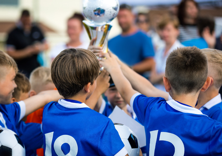Young Soccer Players Holding Trophy. Children Soccer Football Champions. Boys Celebrating Soccer Championship. Winning Team of Sport Kids Tournament. Youth Soccer Cup Winners Parents in the Backgroundの写真素材