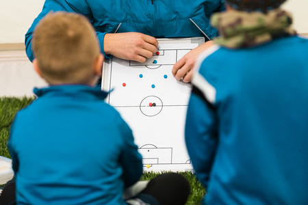 Youth Soccer Coach Coaching Children. Boys Soccer Players Listening Coaches Tactics and Motivational Talk Speech. Teacher Explaining Match Tactics, Players Positions on Soccer Whiteboardの写真素材