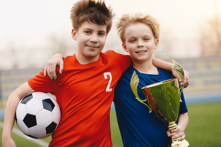Two happy boys soccer players holding soccer ball and golden trophy. Children football players on the field in red and blue jersey sportswearの写真素材