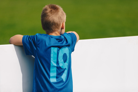 Young Boy in Blue Sports Soccer Jersey Standing Next to the Football Pitch. Soccer Grass on the...