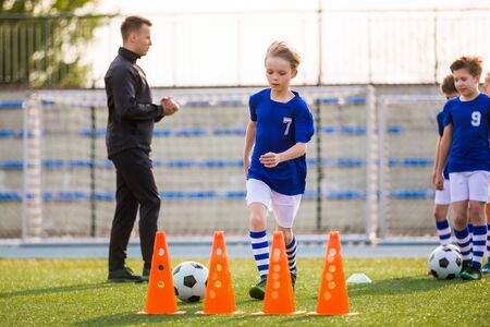 Young Happy Blonde Soccer Player Running with Ball on Training. Coach of Youth Football Team Explaining Drill Exercise in the Background. Caucasian Soccer Players on Practice Sessionの写真素材