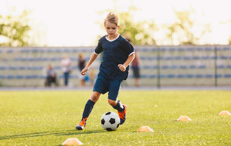 Young boy in blue soccer jersey uniform running after ball on training pitch. Kid improving dribbling skills on practice sessionの写真素材