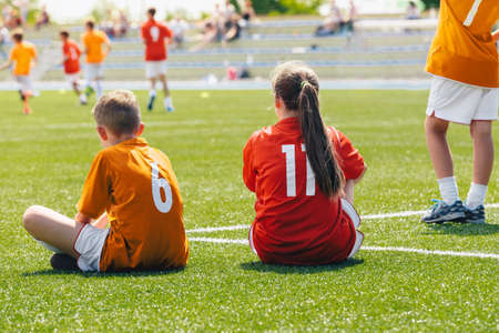 Young boy and girl in a sports team. Happy kids playing sports on summer sunny day. Teammates sitting on grass soccer venue. Young players watching tournament matchの写真素材