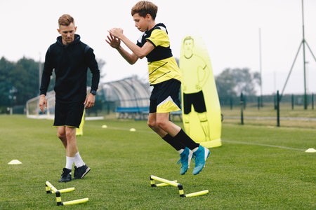 Teenage boy jumping over hurdles. Young soccer athlete practicing strength and agility skills. Boy of youth soccer team on training unit with personal trainer. Soccer coach coaches a young playerの写真素材