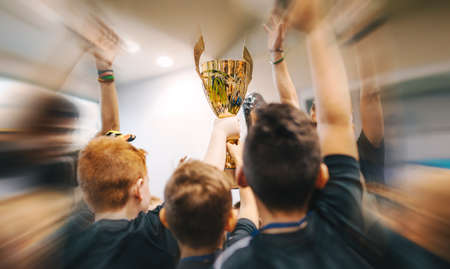 Group of Happy Boys in Sports T-shirts Pick Up the Golden Cup. School Kids Winning Indoor Sport Championship. Sports Tournament Competition For Childrenの写真素材