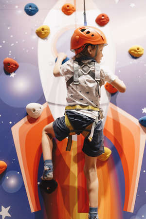 Young boy hanging on rope at indoor climbing wall. Kid in helmet having fun at bouldering wall....