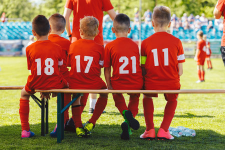 Football players in youth team sitting on wooden bench. Kids in red shirts in school sports team. Boys watching tournament game at soccer stadiumの写真素材