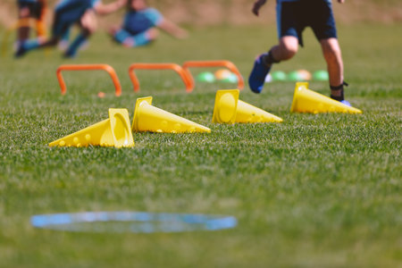 Sports outdoor training trail at grass field. Sports practice summer camp for children. Line of yellow cones and orange hurdles. A young boy attending sports education class at summer campの写真素材