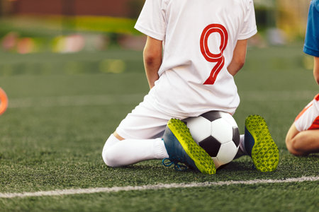 Young caucasian soccer player sitting on soccer at football pitch. Little boys on sports training watching training game. Children in soccer jerseyの写真素材