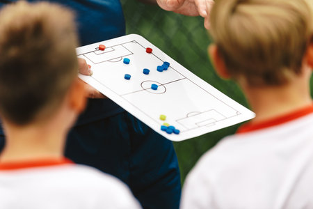 A young coach explains to a group of children the soccer team strategy using a tactics board. School coach coaching kids in a sports team. Strategy board for soccer tactics educationの写真素材