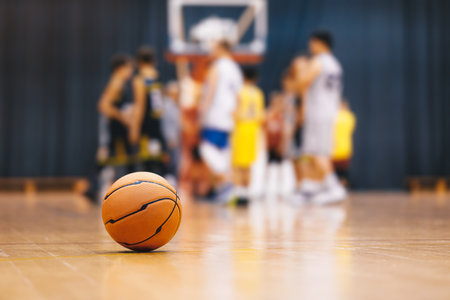 Basketball ball on wooden court. Young boys play basketball match in blurred background. Junior level sports team compete in indoor gameの写真素材