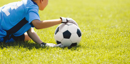 A young boy as a soccer goalie catching a soccer ball during a tournament game. A school football goalkeeper in a blue jersey shirt and sports gloves throwing the ball and playing a football matchの写真素材