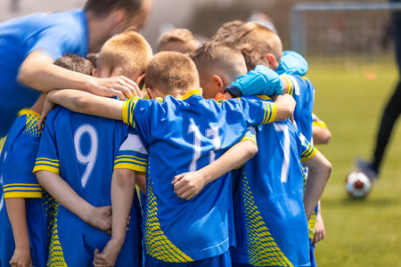 The soccer team huddles in a circle with a coach during the time break. Kids on the football team stand united and listen to the coach's motivational speech. Sports children in blue soccer uniformsの写真素材