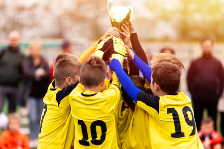Happy children in sports team celebrating victory. Kids winning the game and rising golden cup. Boys football team huddling in circle and jumping in happiness. Youth sports team succeed in tournamentの写真素材