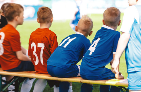 Football soccer match for children. Boys play sports outdoors. Kids are waiting on a bench. Soccer players in red and blue soccer jerseysの写真素材