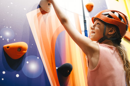 Indoor climbing class for kids. School girl having fun in indoor playground for children. Happy little girl in red helmet climbing the wall in bouldering centerの写真素材