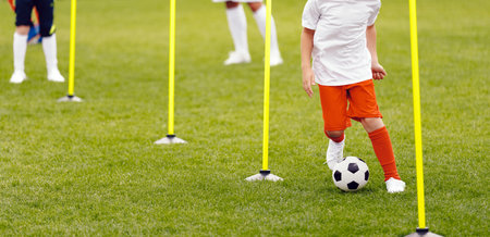 Young athlete running with soccer ball in slalom between agility poles. Soccer education for kids. Training with agility poles. Football players on a drill. Football training for junior footballersの写真素材