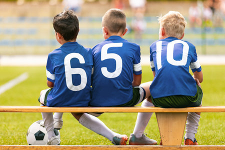 Boys in blue soccer jerseys with white numbers sit on a wooden substitution bench, watching the game, united as a teamの写真素材