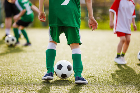 Children in soccer uniforms play on a sunny field. A kid in green stands over a soccer ball, with teammates and opponents in the background practicing and kicking soccer ballsの写真素材