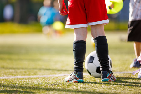 A young soccer player in red shorts and black socks stands on a field with a soccer ball, ready to play. Other kids and practice gear are visible in the blurred backgroundの写真素材
