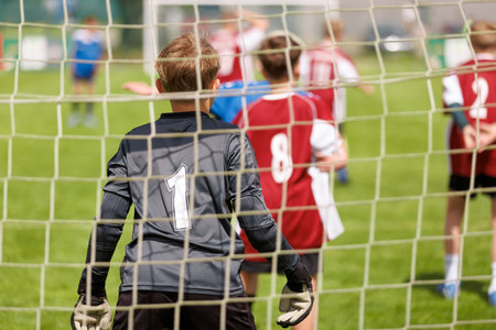 Football Goals In Action. Youth Soccer Goalkeeper Focused on the Game from Behind the Net During a Match on a Sunny Day. Young Boys Play as Soccer Football Goalieの写真素材