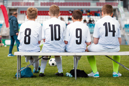 Youth Soccer Players on the Bench Waiting for Substitution. Junior Football Team in Uniform During Match. Kids Preparing for Soccer Gameの写真素材