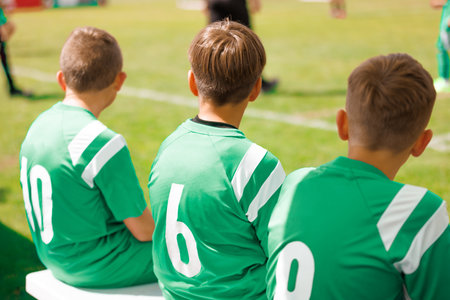 Boys in Green Jerseys Sitting on Sideline Bench. Boys Soccer Team Sitting on Bench. Young Players in Green Jerseys Watching Football Game during Youth Sports Eventの写真素材