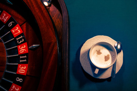 A roulette wheel with red and black numbered pockets stands next to a cup of coffee on a table,...